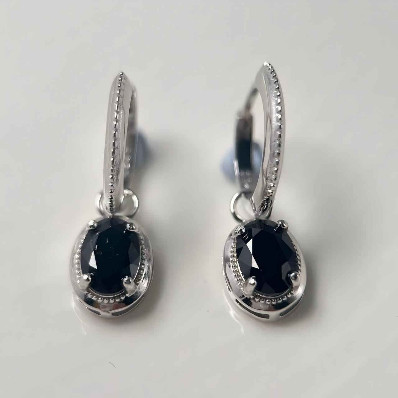 A pair of silver-toned hoop earrings with oval black faceted gemstones, displayed on a light gray reflective surface.