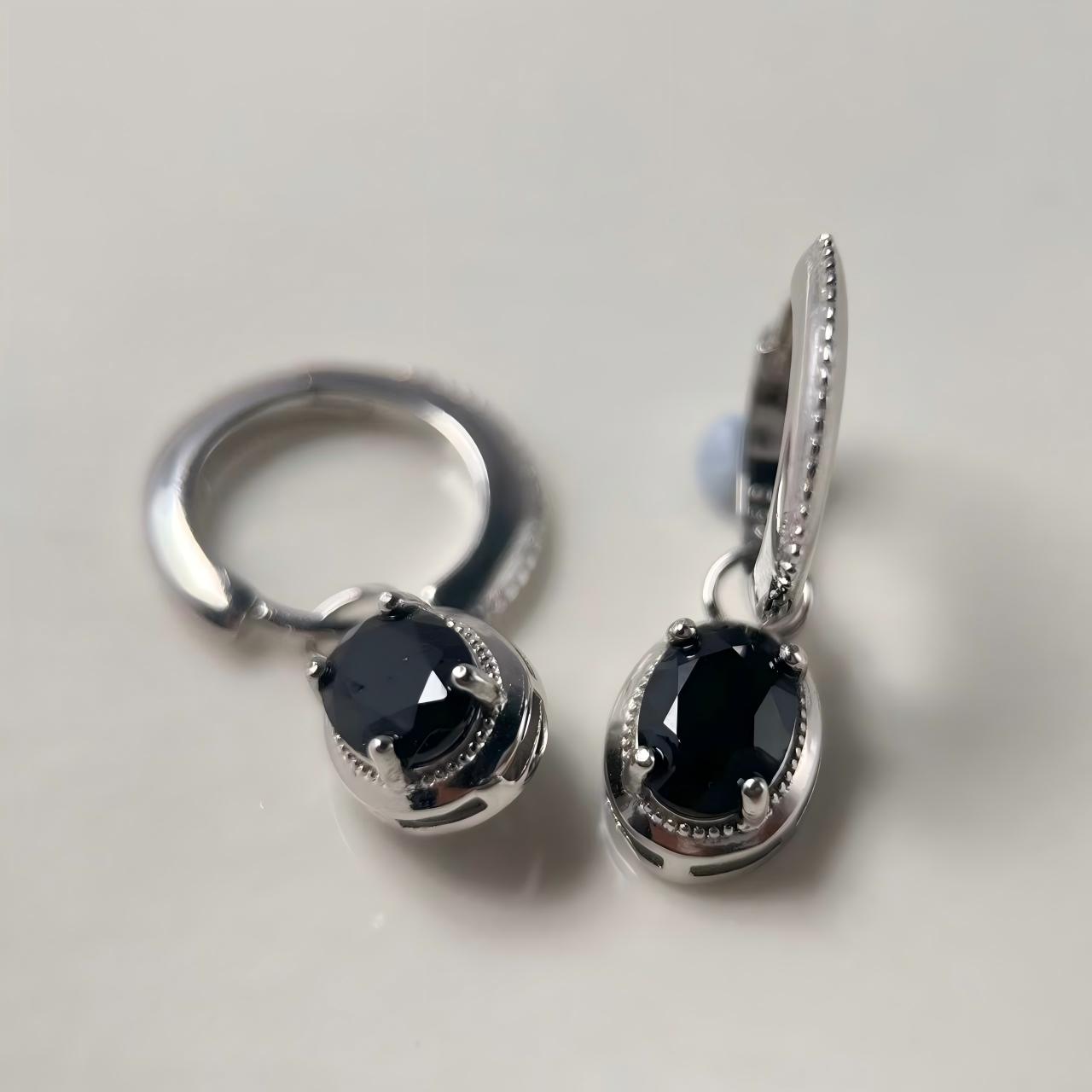 A pair of silver-toned huggie hoop earrings with oval black gemstones, displayed on a light gray background.