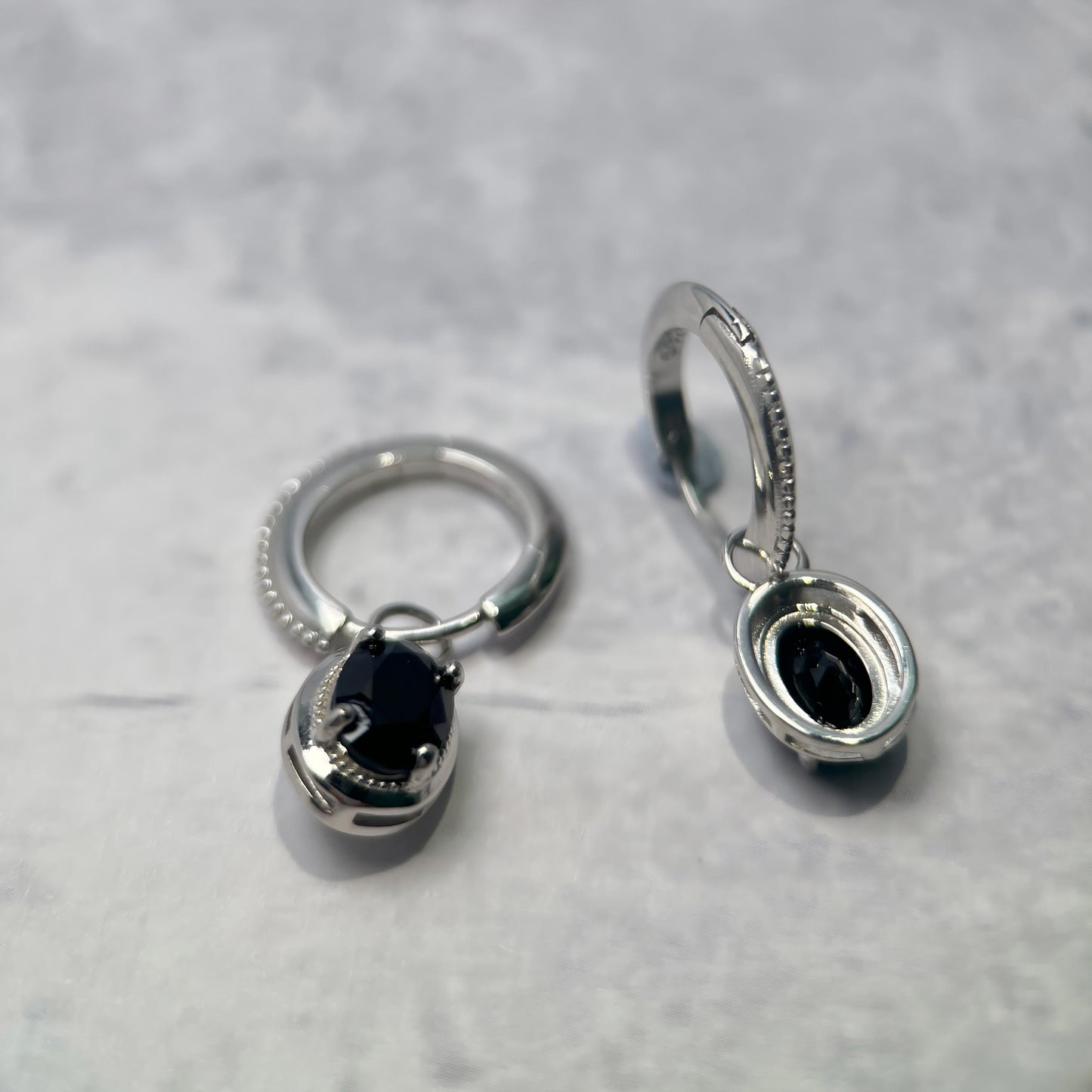 A pair of silver-toned hoop earrings with oval black stone inlays, displayed on a light gray textured surface.