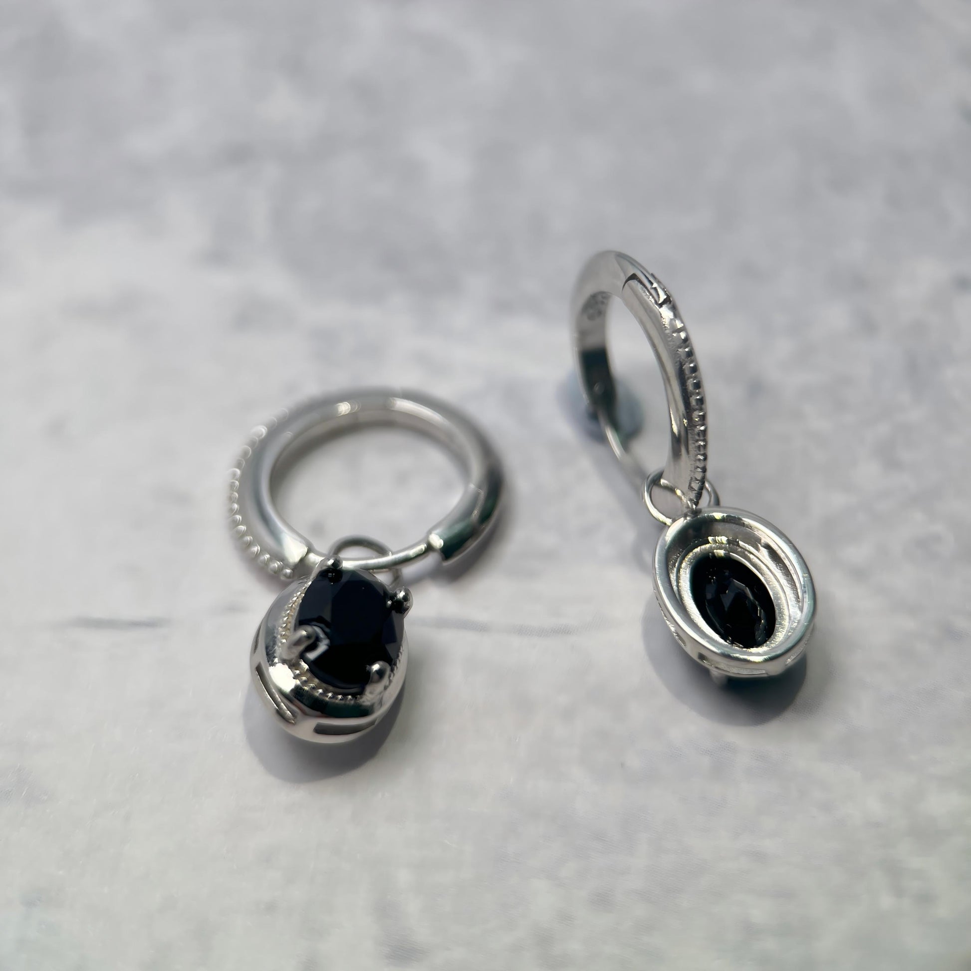 A pair of silver-toned hoop earrings with oval black stone inlays, displayed on a light gray textured surface.