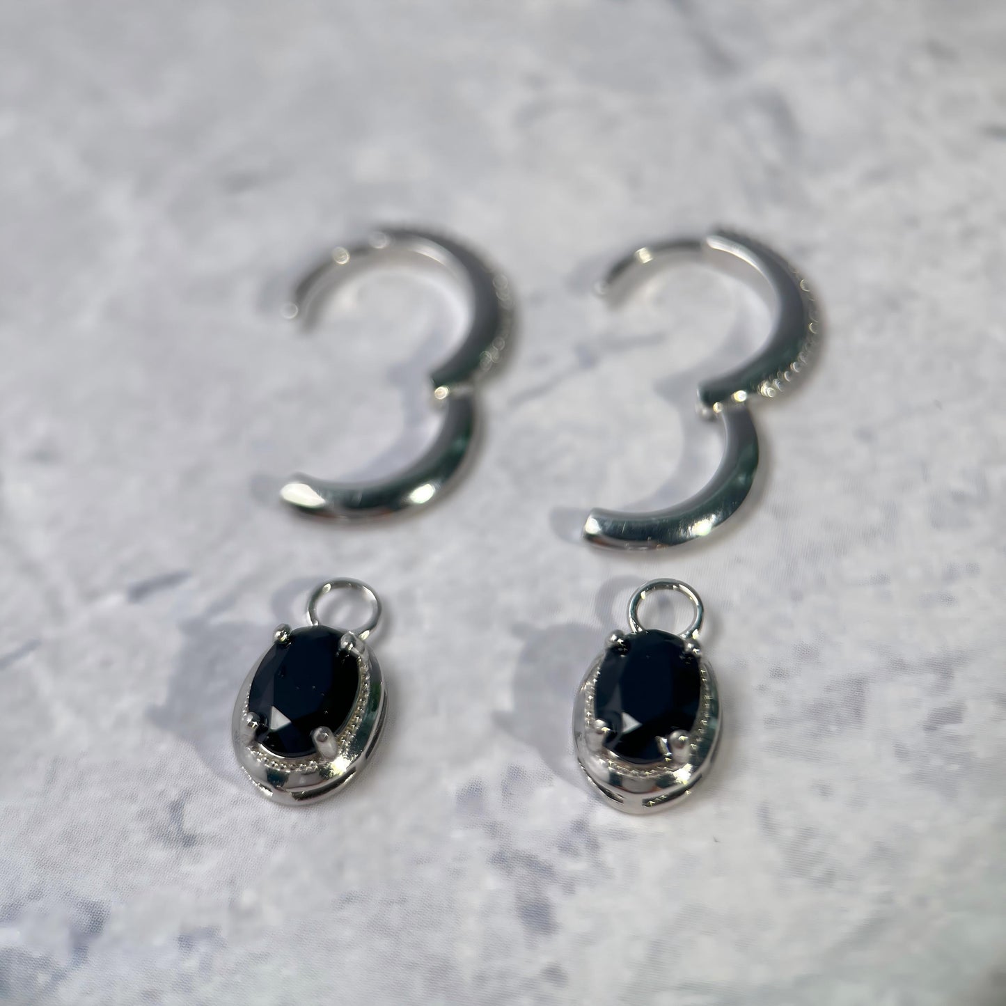 A pair of silver hoop earrings with matching oval drop charms featuring black faceted gemstones, displayed on a light gray textured surface.