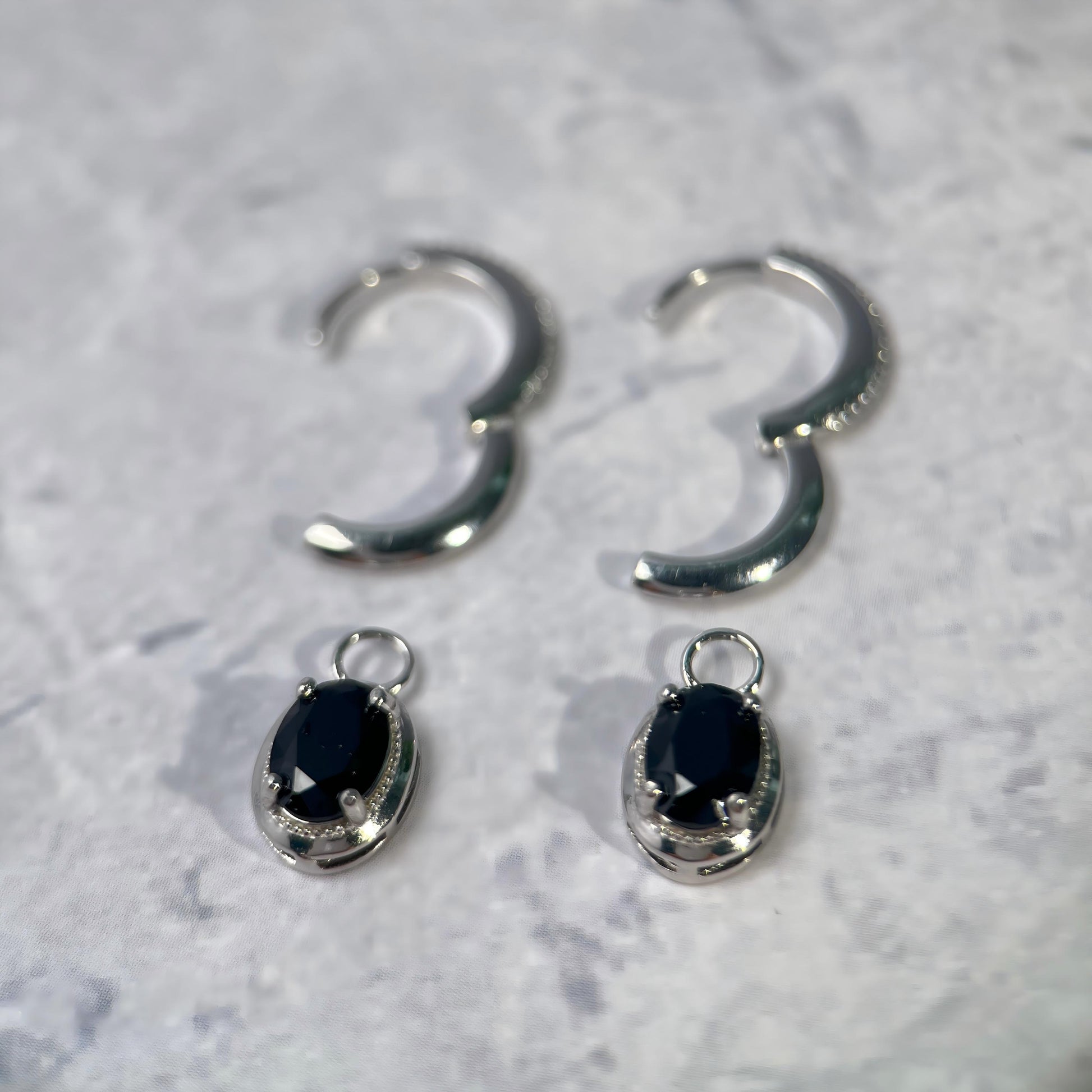 A pair of silver hoop earrings with matching oval drop charms featuring black faceted gemstones, displayed on a light gray textured surface.