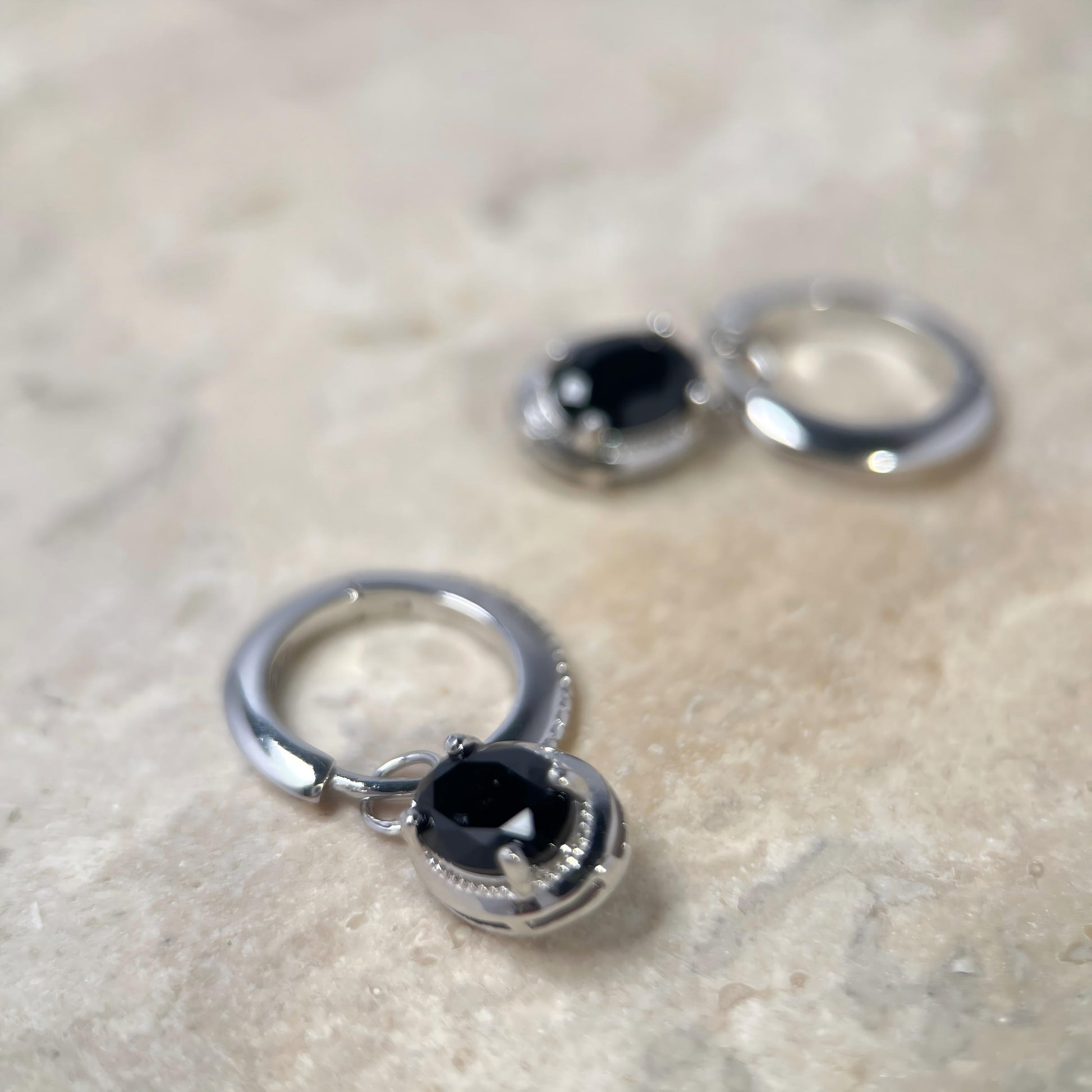 A pair of silver-toned hoop earrings with oval black gemstones, displayed on a light beige stone surface.