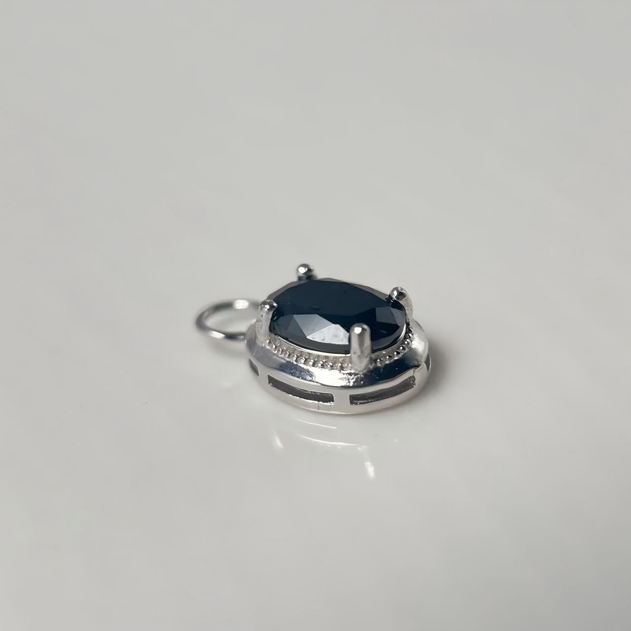 A silver-toned oval pendant charm featuring a faceted black gemstone, set in a polished silver bezel with a small attached loop at the top, photographed on a neutral light gray background.