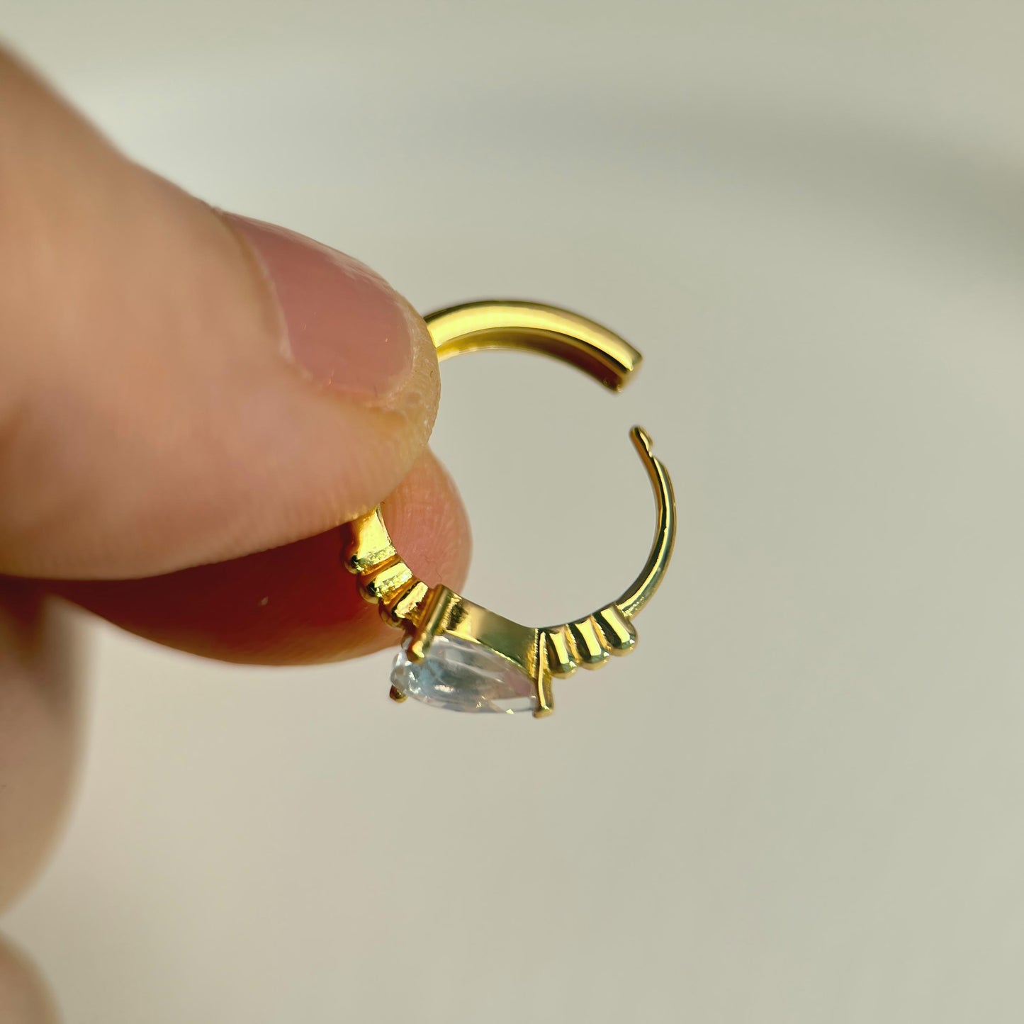 Gold hoop earring with a clear gemstone held between fingers against a neutral background